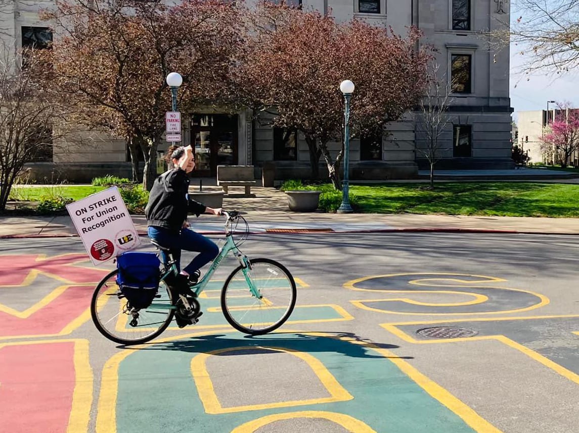 Woman on bicycle riding from left to right.The signs say "On strike for union recognition."