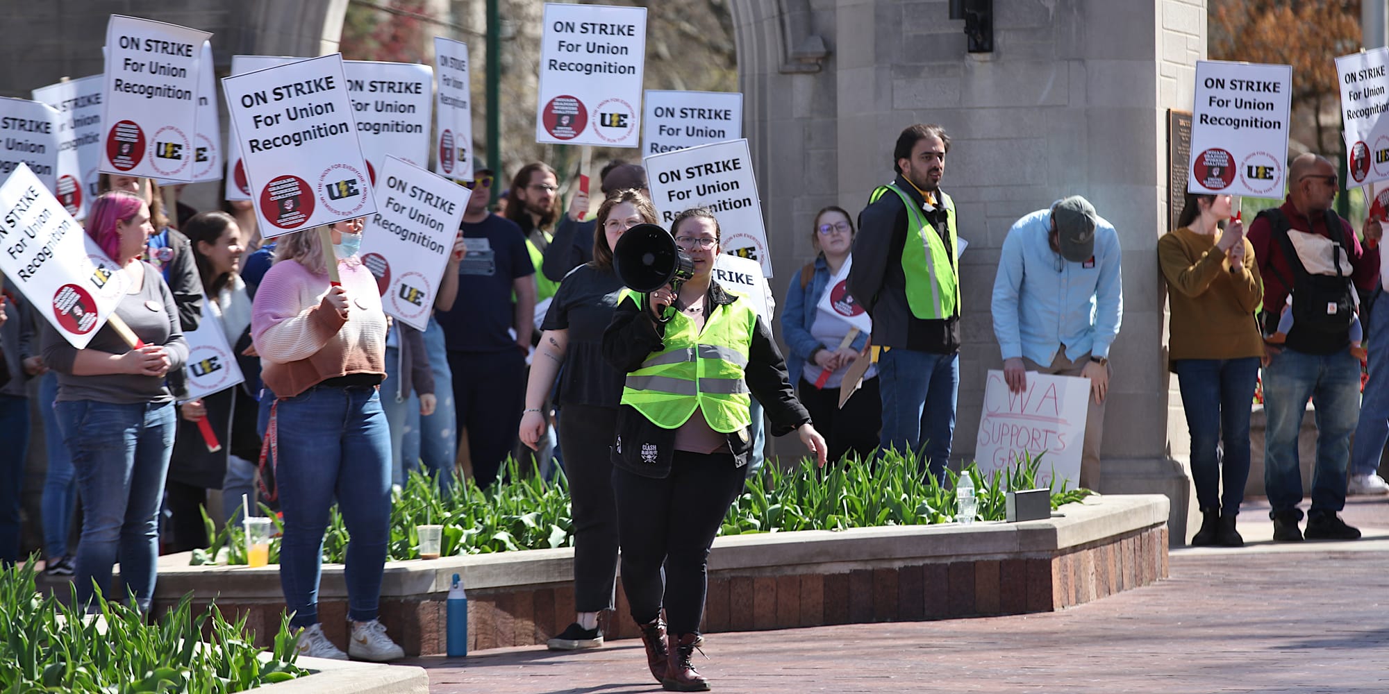 Woman standing in front of a large group of people holding signs. The signs say "On strike for union recognition."