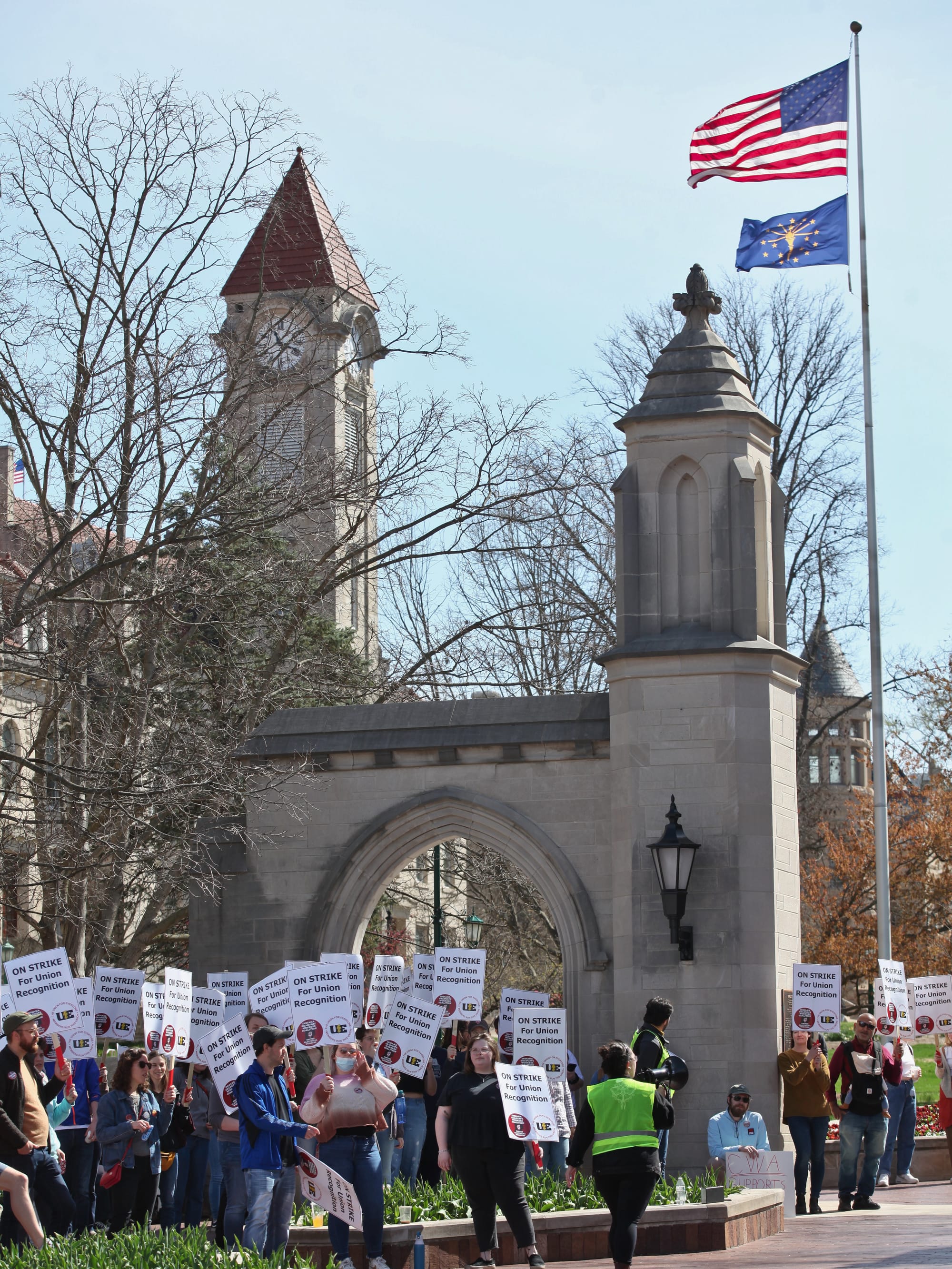 Long shot of group of people standing in front of university gateway holding signs Woman in yellow reflective vest standing in the middle of a crowd of people with signs. The signs say:"On strike for union recognition."