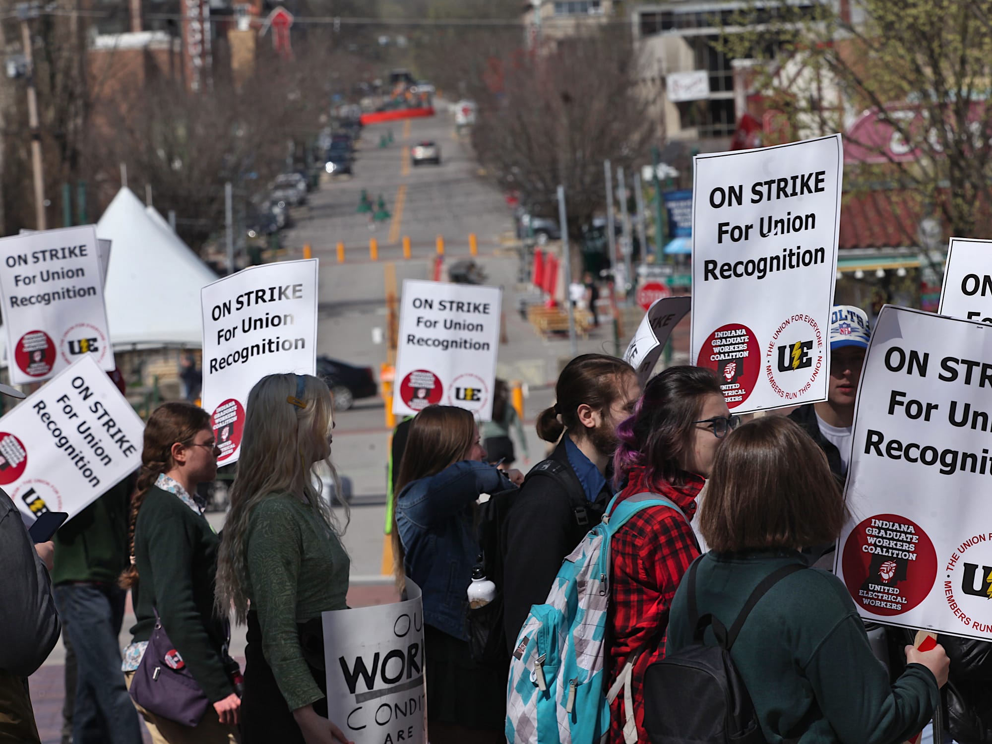 View down street with people in the foreground holding signs. The signs say "On strike for union recognition."