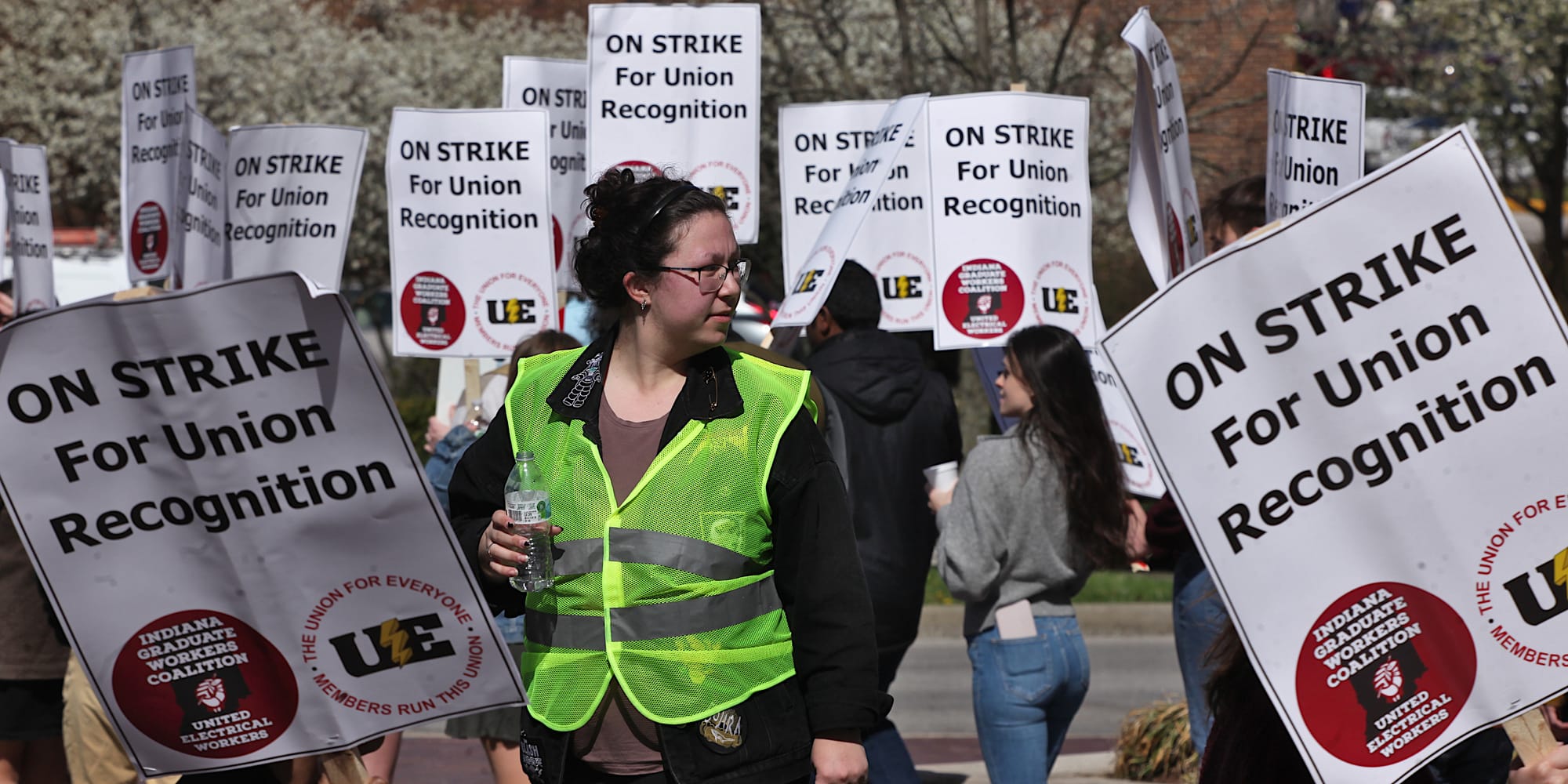 Woman in yellow reflective vest standing in the middle of a crowd of people with signs. The signs say:"On strike for union recognition."