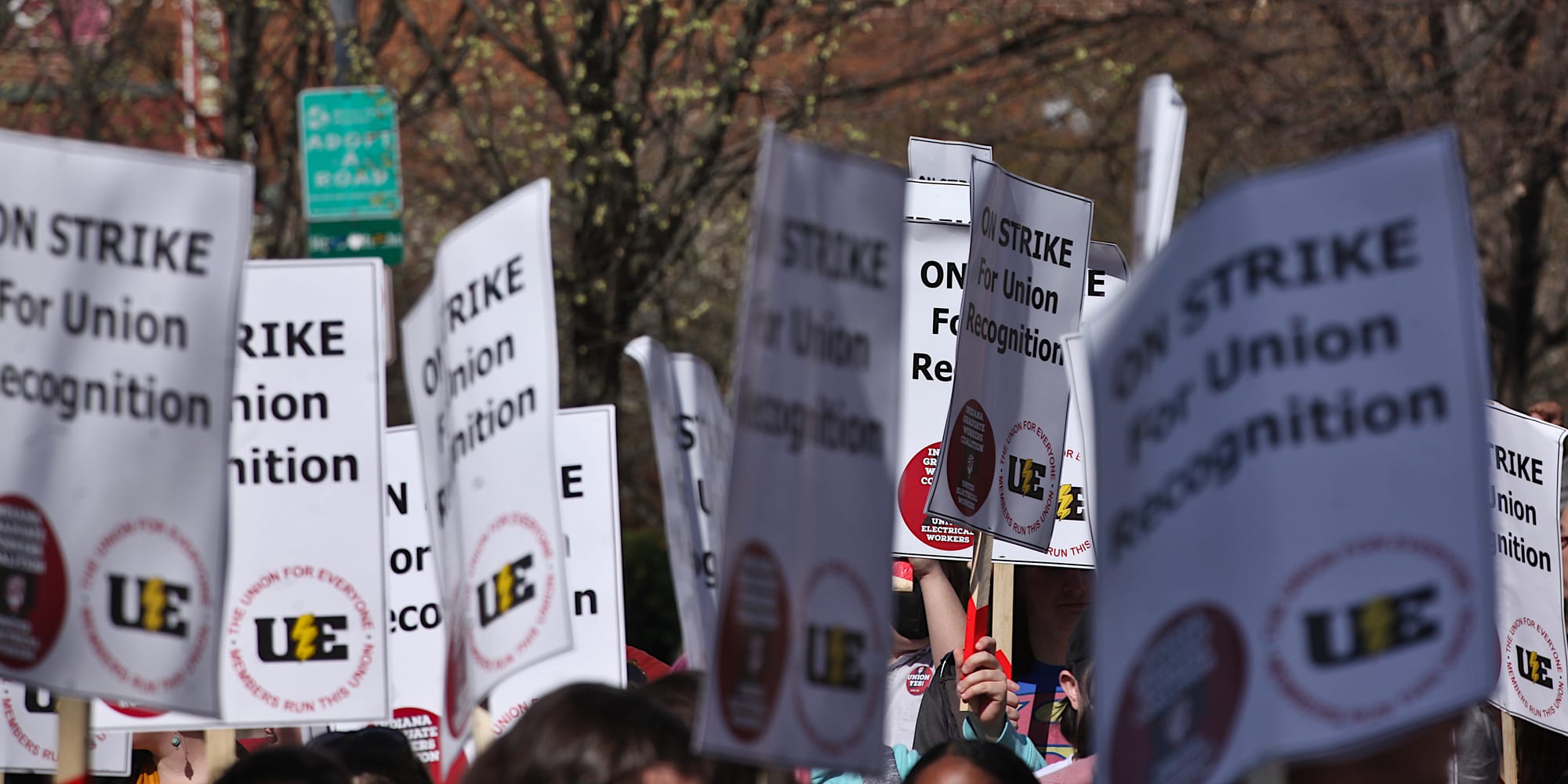 Group of people holding signs. Woman in yellow reflective vest standing in the middle of a crowd of people with signs. The signs say:"On strike for union recognition."