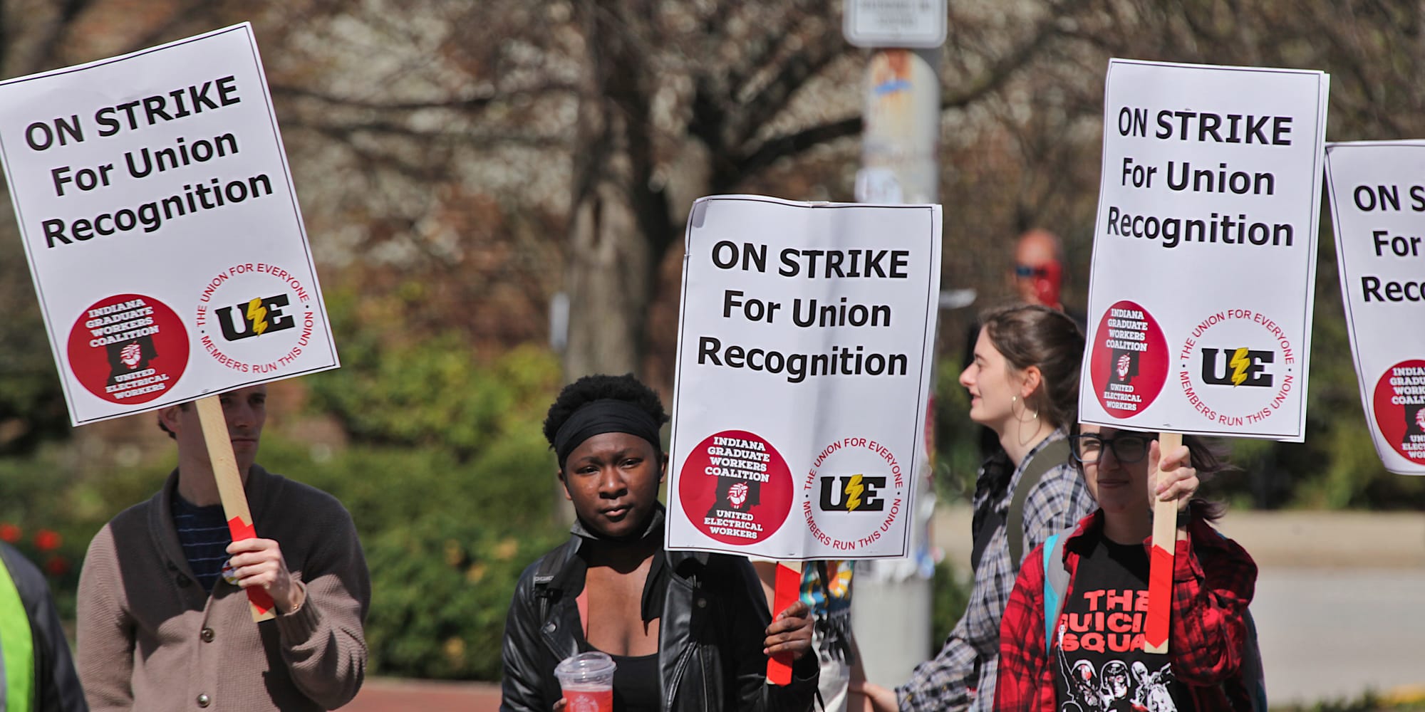 People holding signs. Long shot of group of people standing in front of university gateway holding signs Woman in yellow reflective vest standing in the middle of a crowd of people with signs. The signs say:"On strike for union recognition."