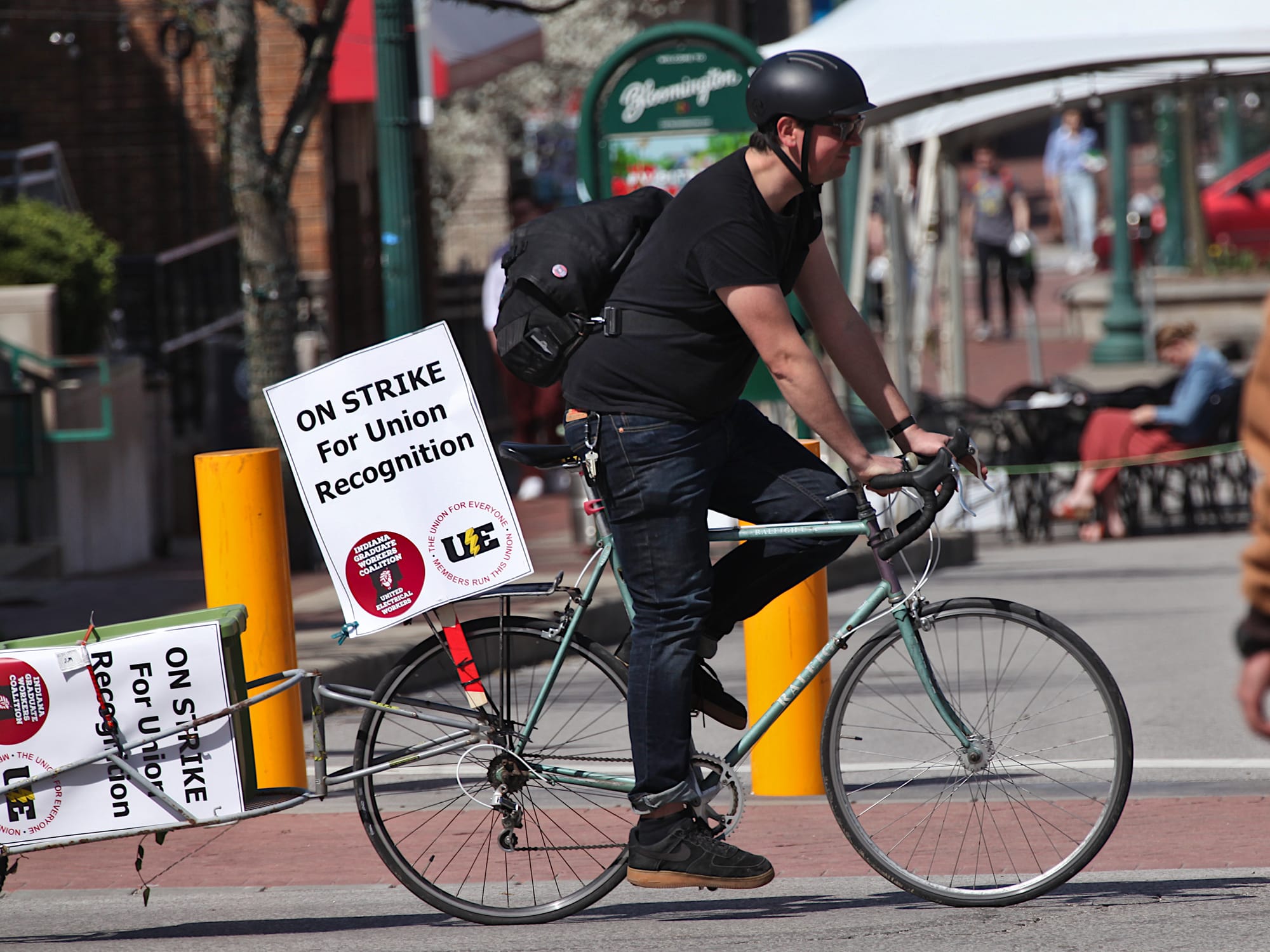 Man riding a bicycle from left to right. A sign is affixed to the back of the bicycle and to a trailer. The signs say "On strike for union recognition."