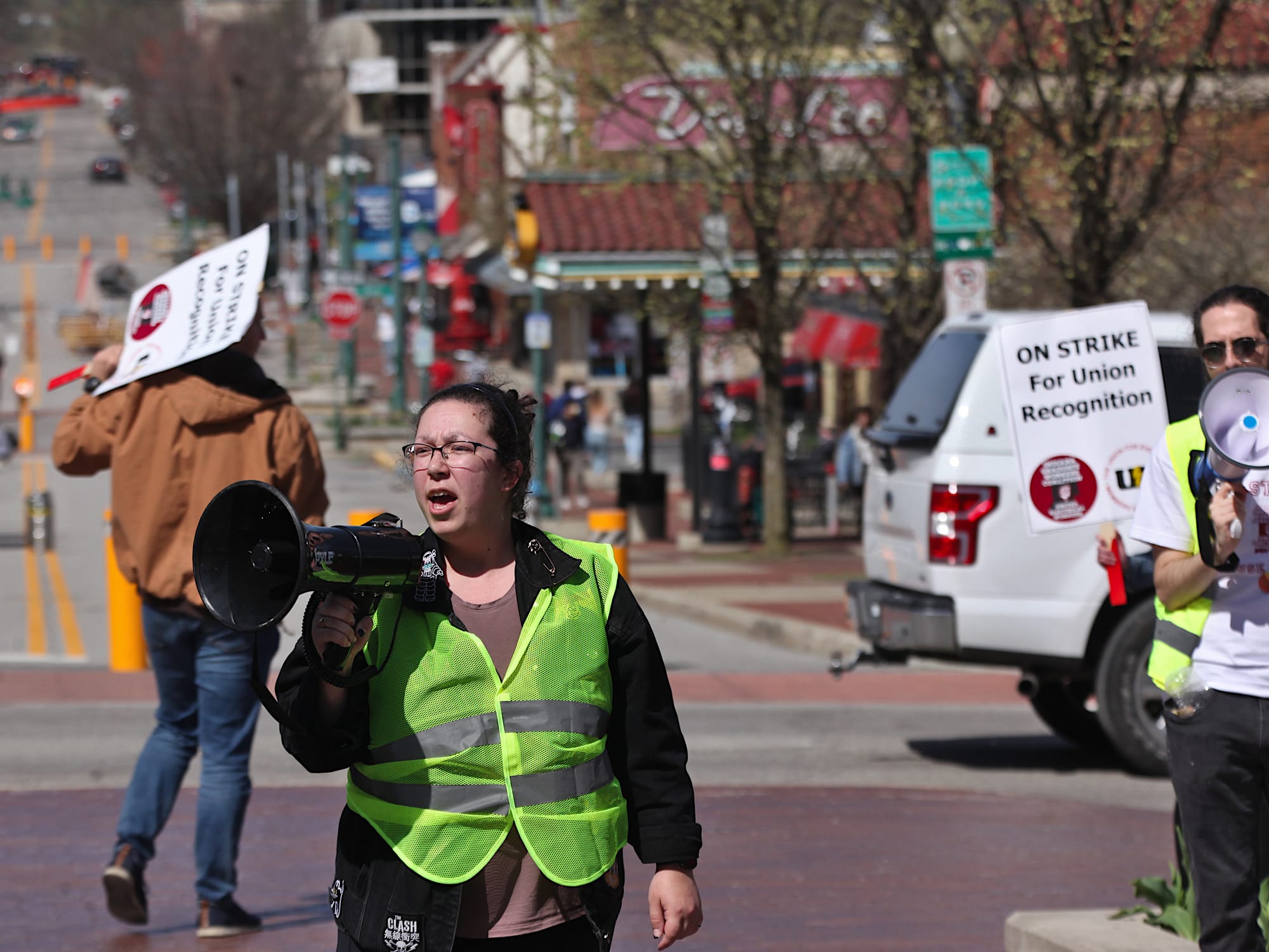 Woman speaking into a megaphone surrounded by people holding signs. The signs say "On strike for union recognition."