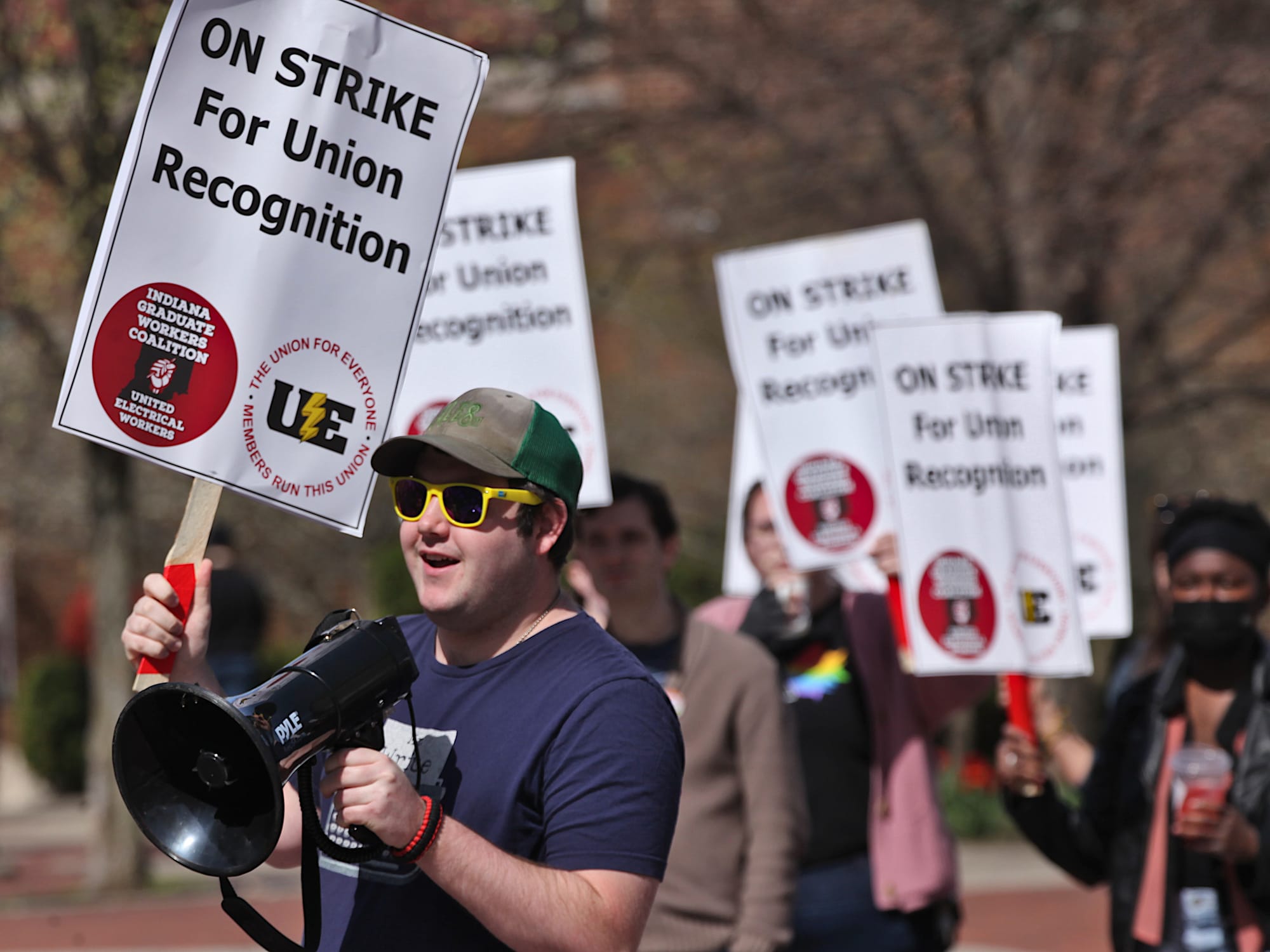 Man speaking into a microphone. People holding signs in the background. The signs say "On strike for union recognition."