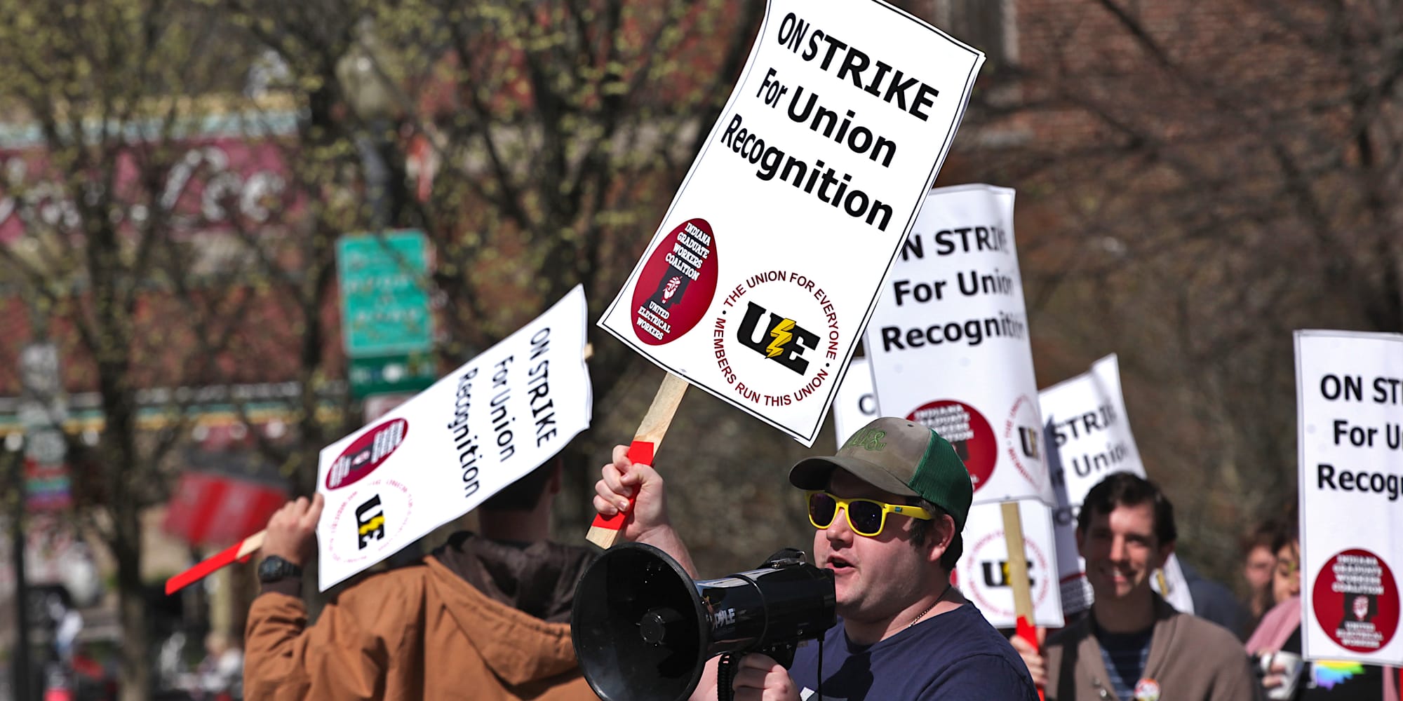 Man holding a sign among others with signs. Woman in yellow reflective vest standing in the middle of a crowd of people with signs. The signs say:"On strike for union recognition."