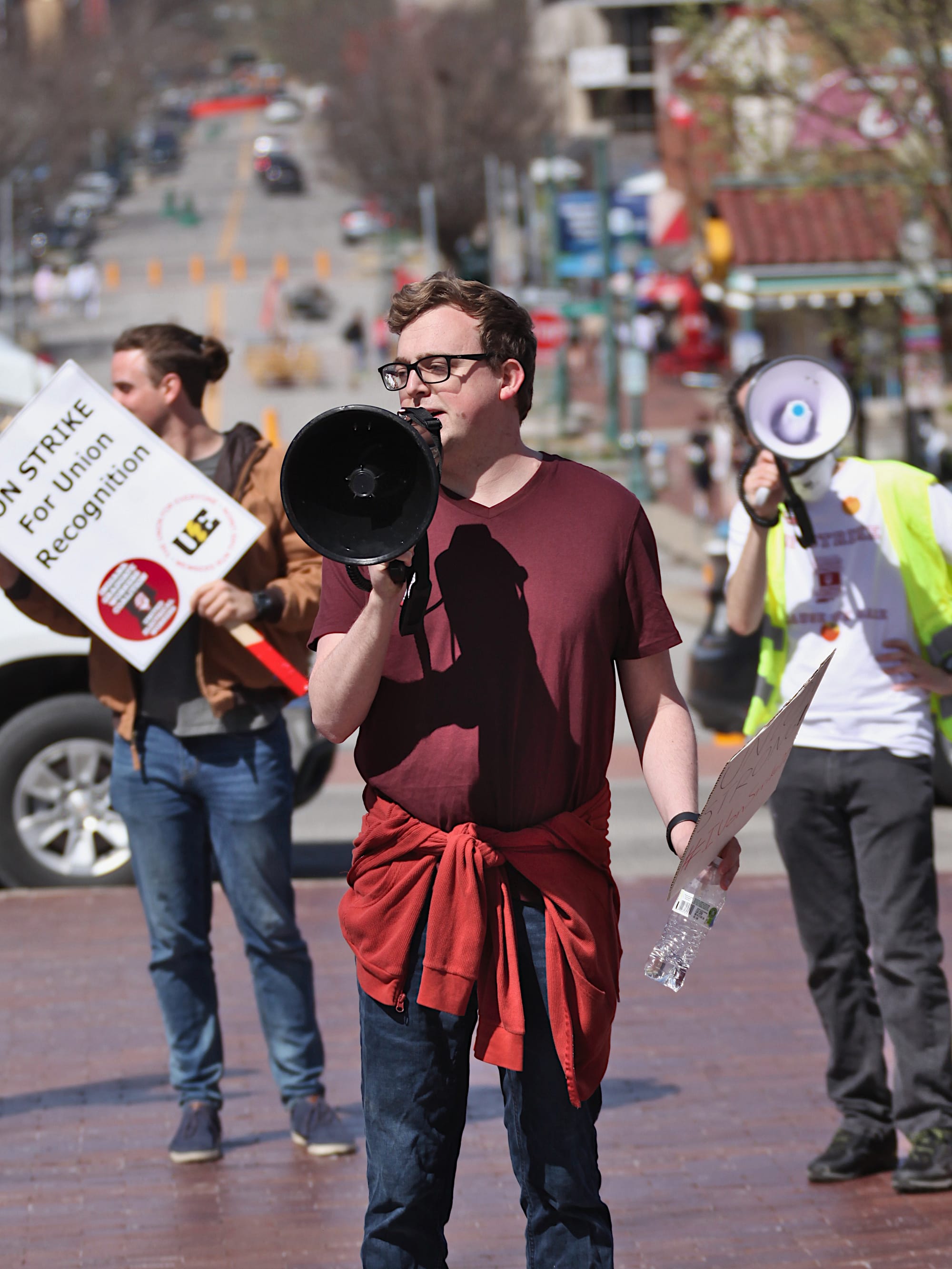 Man speaking into a microphone. People holding signs in the background. The signs say "On strike for union recognition."