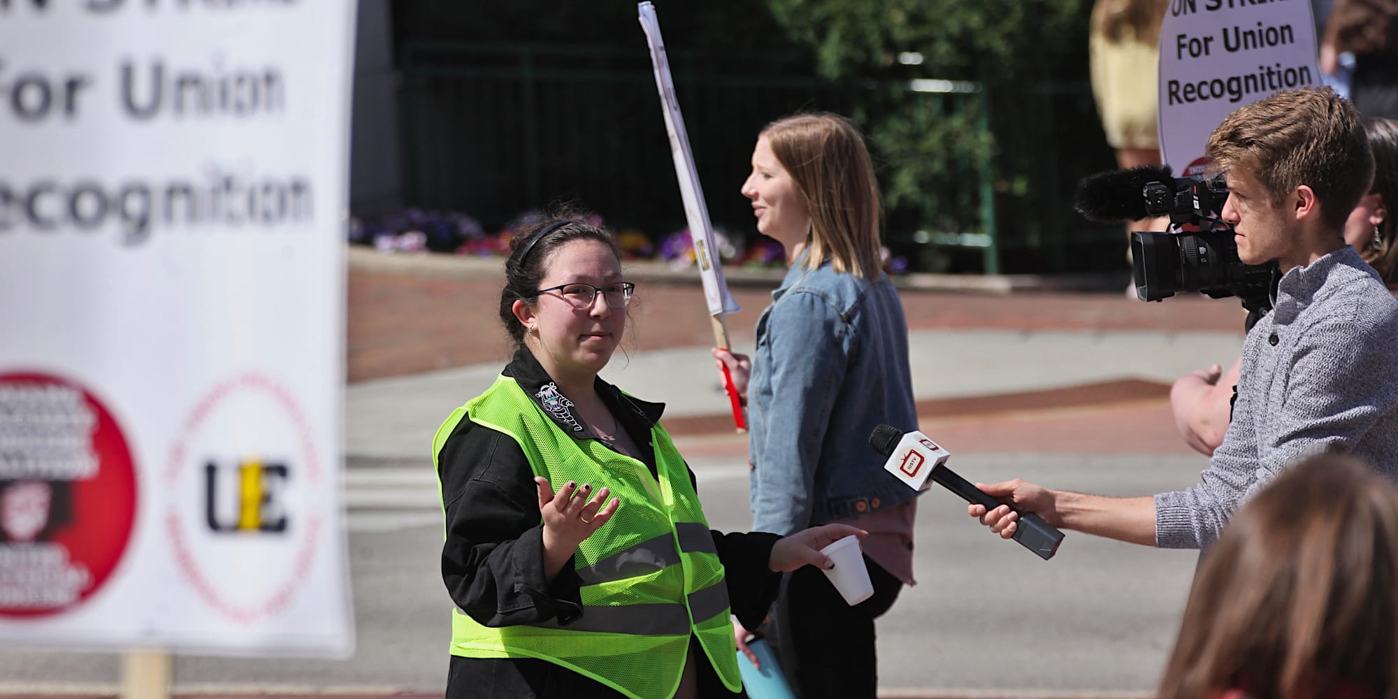 Woman in yellow vest getting interviewed by a man holding a microphone towards here. People are marching past with signs. The signs say "On strike for union recognition."