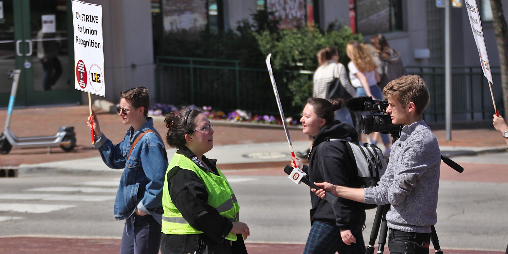 Woman in yellow vest getting interviewed by a man holding a microphone towards here. People are marching past with signs. The signs say "On strike for union recognition."