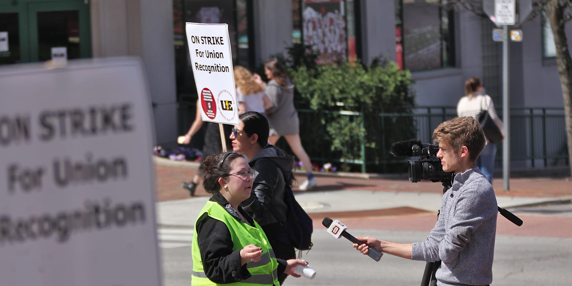 Woman in yellow vest getting interviewed by a man holding a microphone towards here. People are marching past with signs. The signs say "On strike for union recognition."