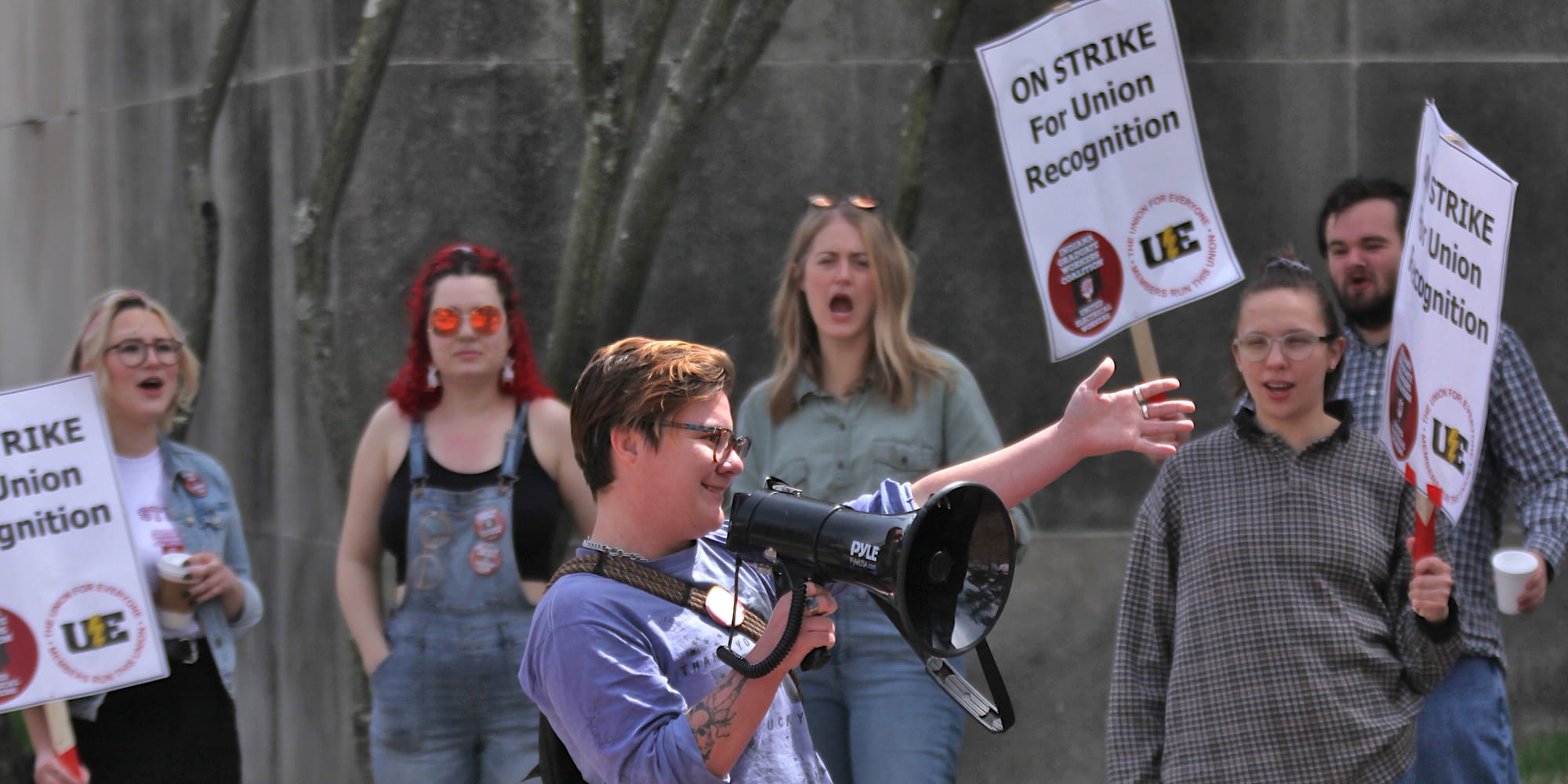 Woman with megaphone speaking into it. People holding signs in the background.The signs say "On strike for union recognition."