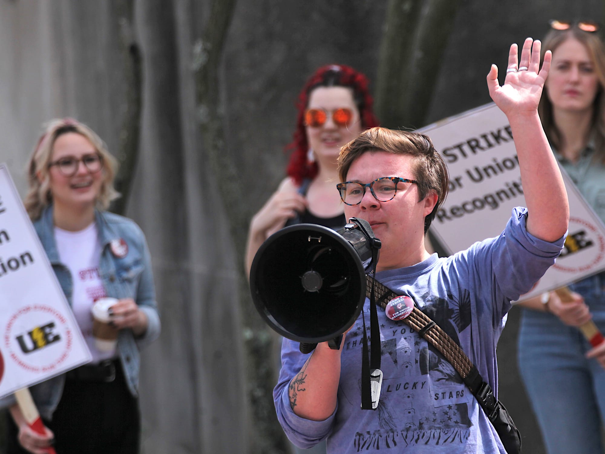 Woman with megaphone speaking into it. People holding signs in the background. The signs say "On strike for union recognition."