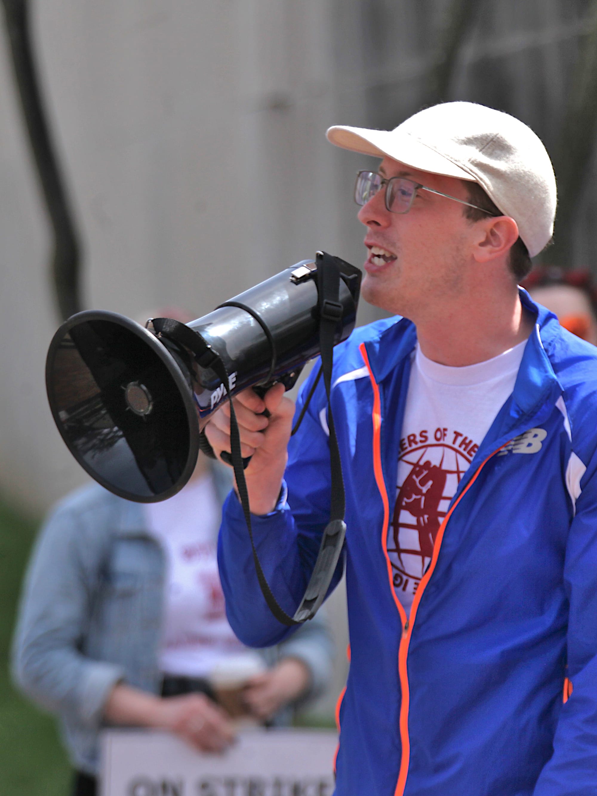 Man with megaphone shouting into it. People holding signs in background. The signs say "On strike for union recognition."