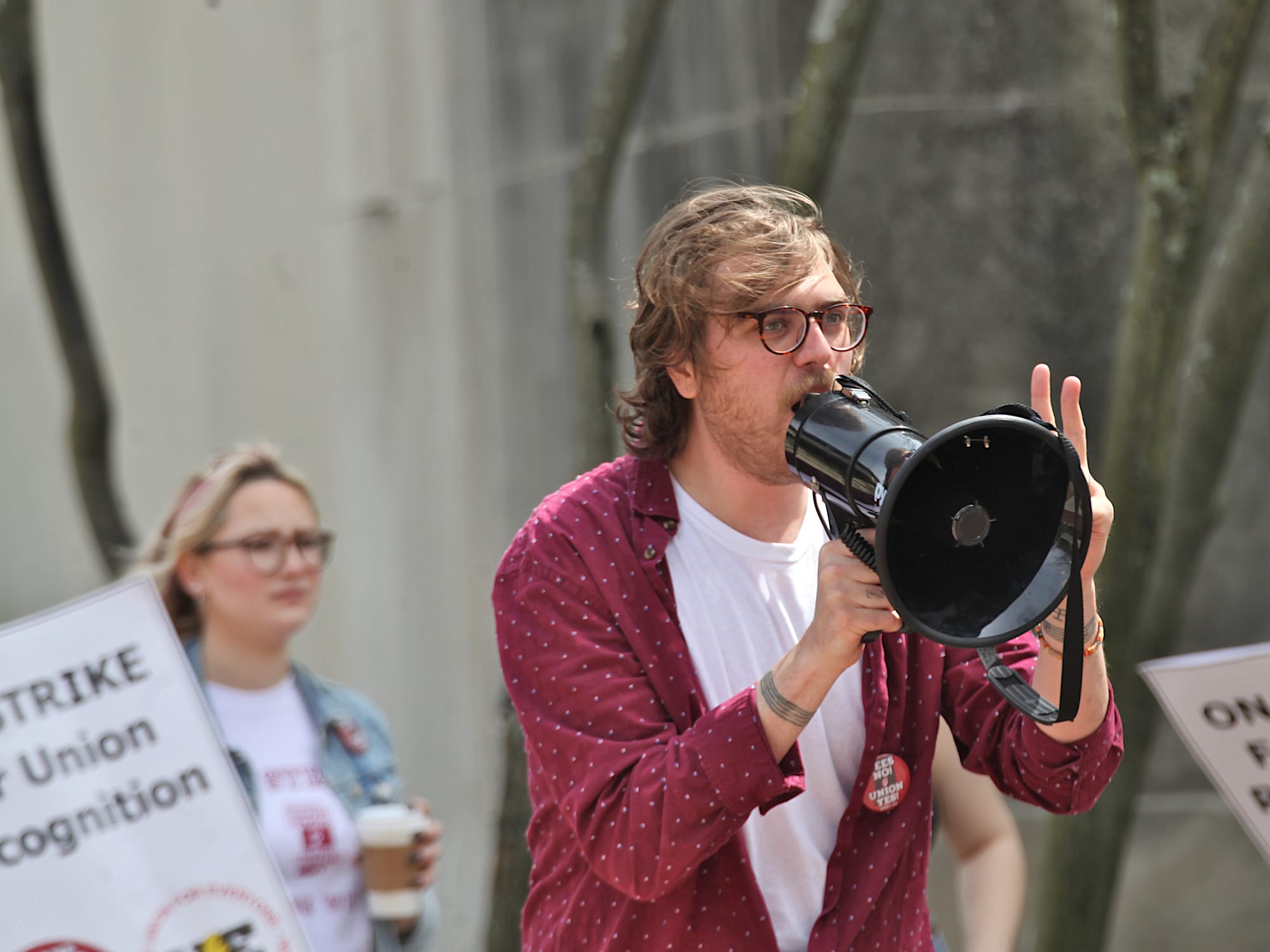 man with megaphone speaking into it. He's standing in front of a crowd with signs that say "On strike for union recognition."