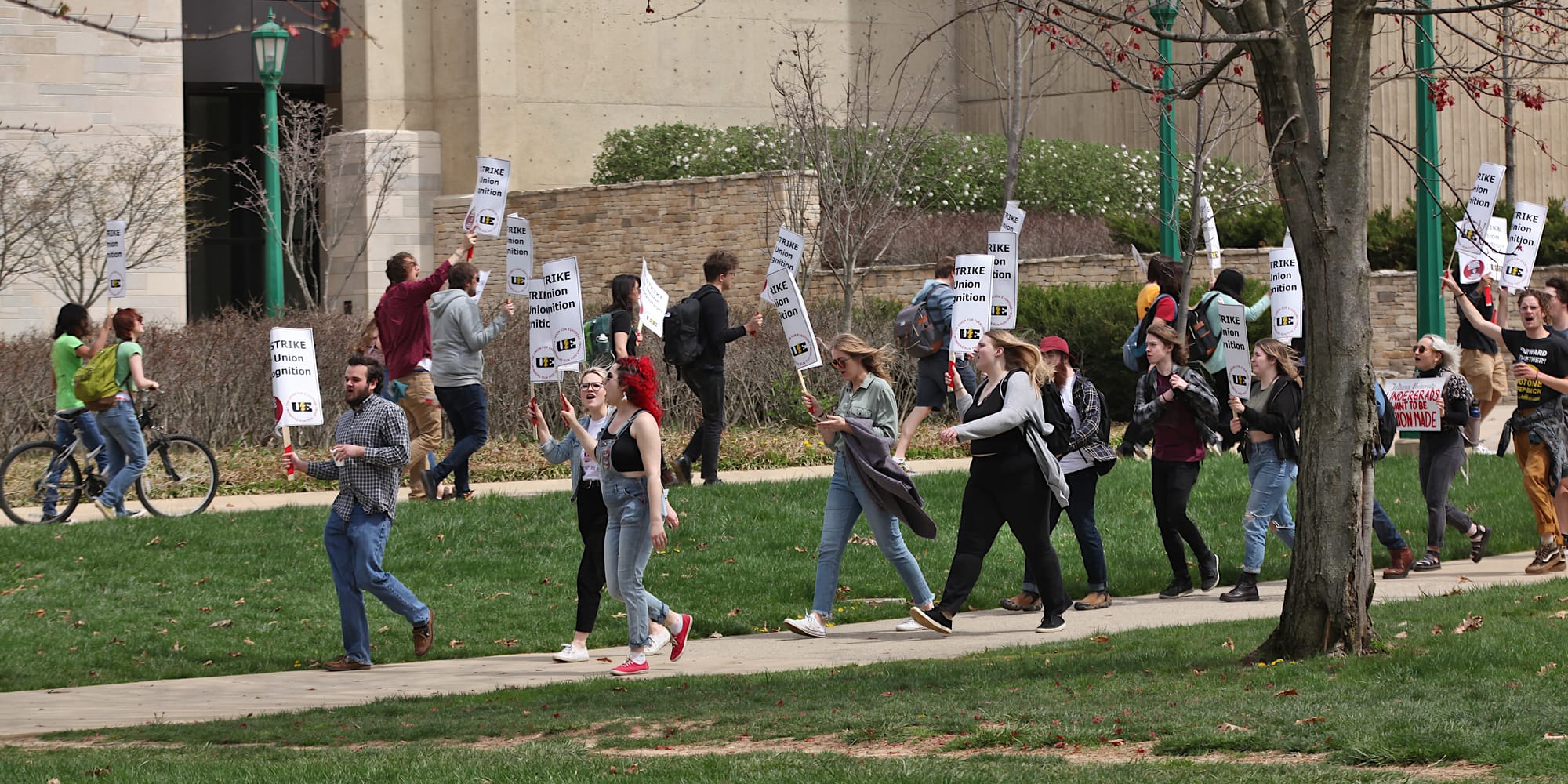 A long shot of a group of people walking along a sidewalk that loops around so that some people are facing to the right and others to the left. The signs say "On strike for union recognition."