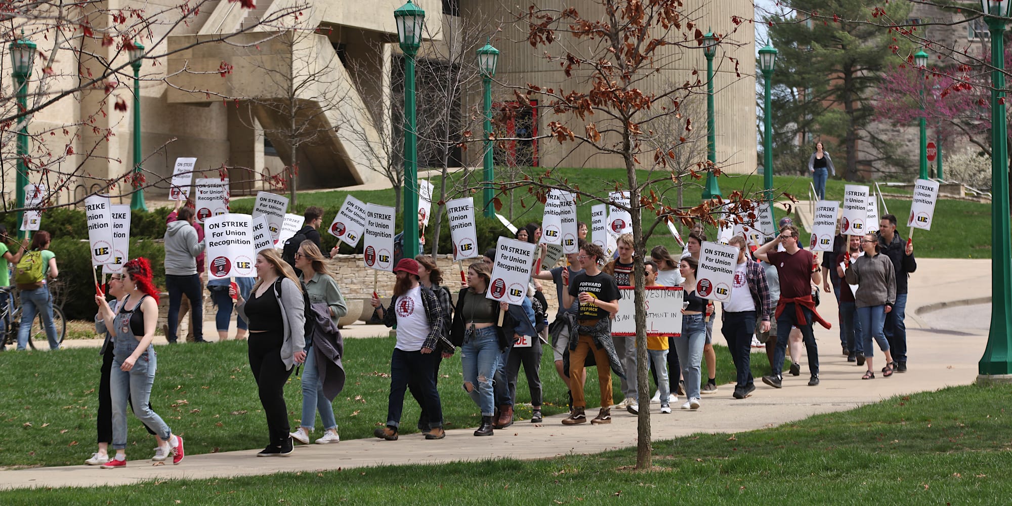 A long shot of a group of people walking along a sidewalk that loops around so that some people are facing to the right and others to the left. The signs say:"On strike for union recognition."