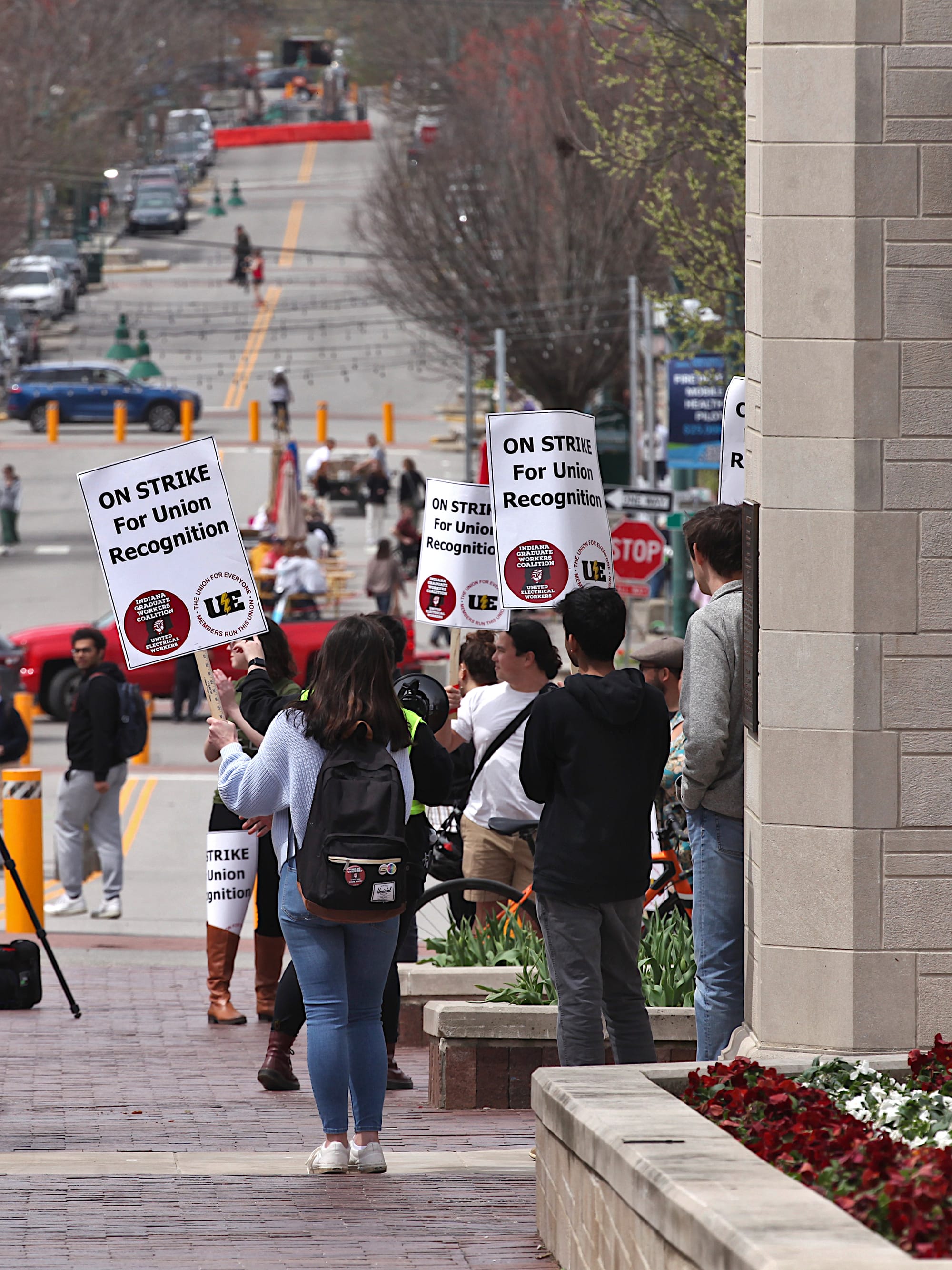 View down street with group of people on right of frame holding signs. The signs say "On strike for union recognition."