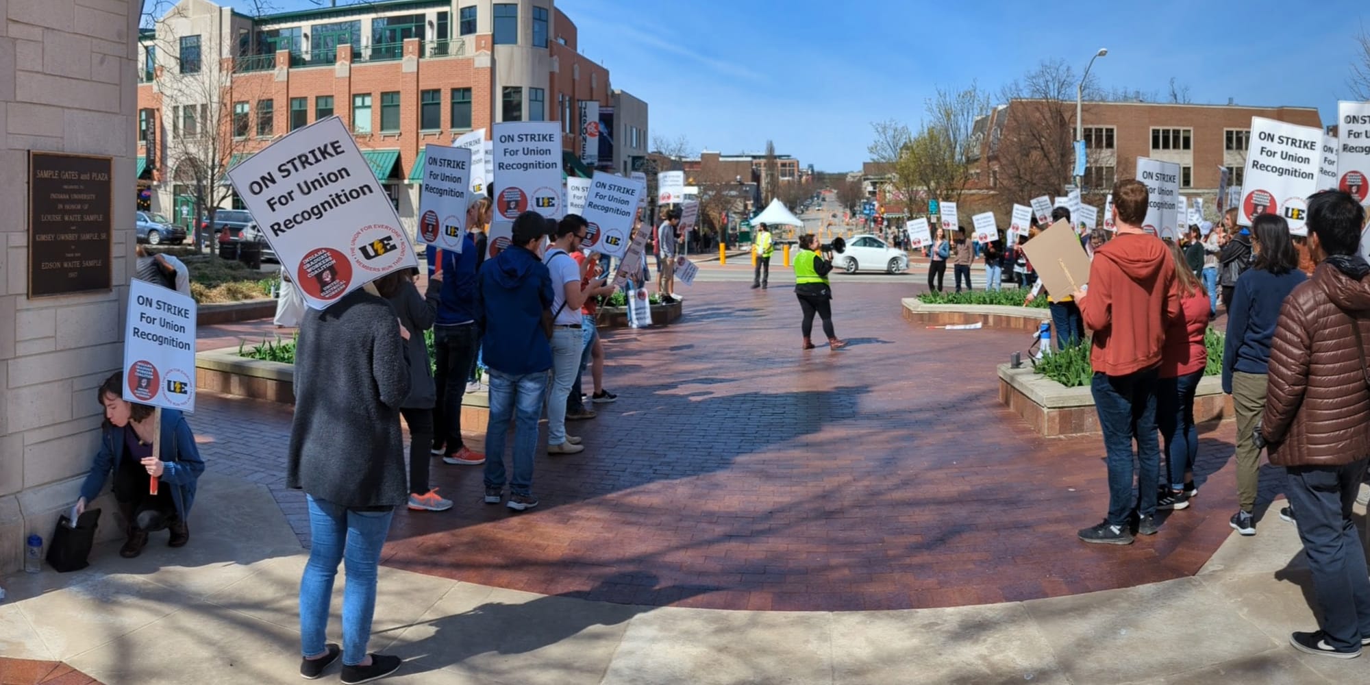 long shot down street with people on the left of the frame holding signs. Long shot of group of people standing in front of university gateway holding signs Woman in yellow reflective vest standing in the middle of a crowd of people with signs. The signs say:"On strike for union recognition."