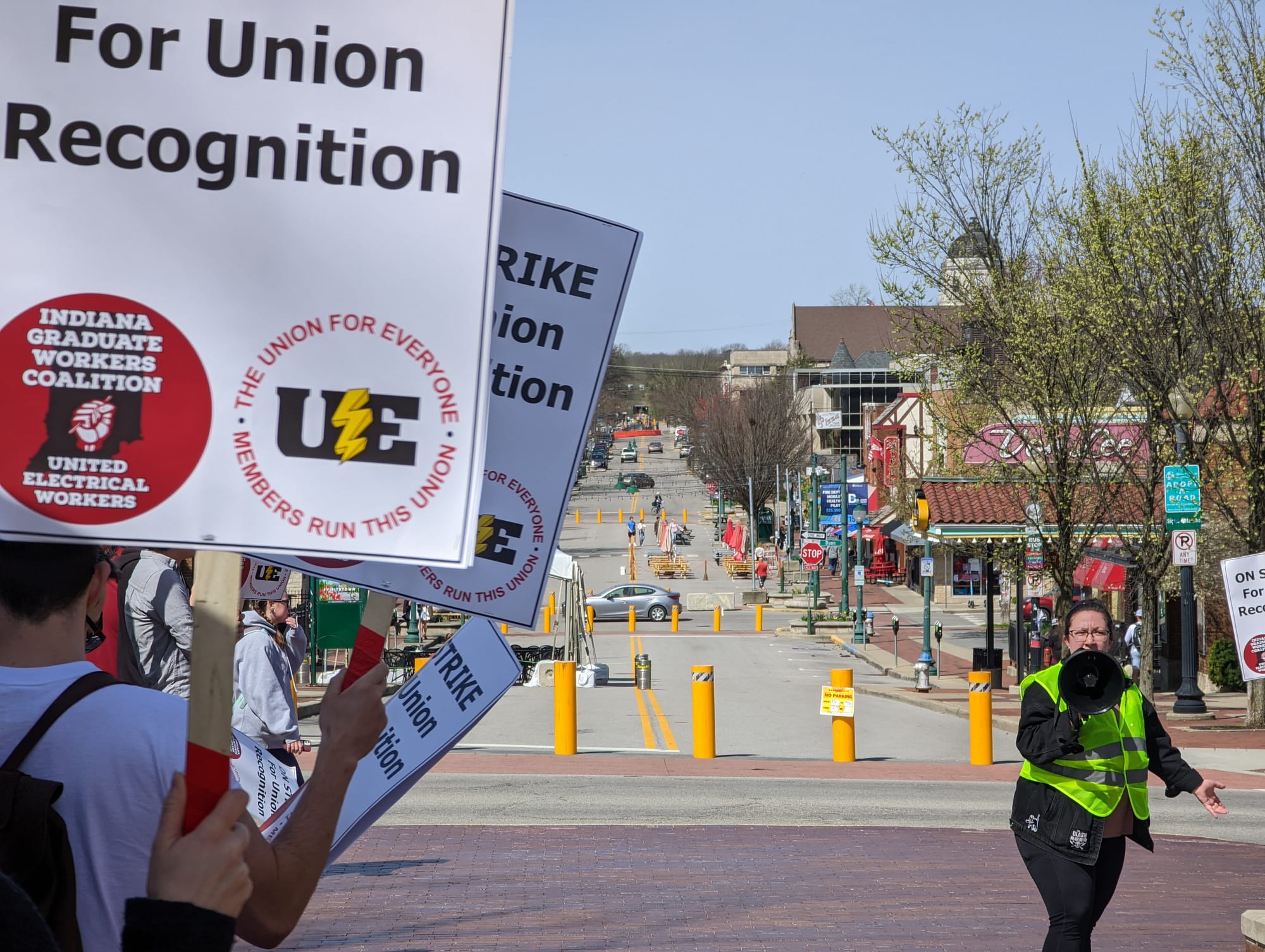 long shot down street with people on the left of the frame holding signs. Long shot of group of people standing in front of university gateway holding signs Woman in yellow reflective vest standing in the middle of a crowd of people with signs. The signs say:"On strike for union recognition."