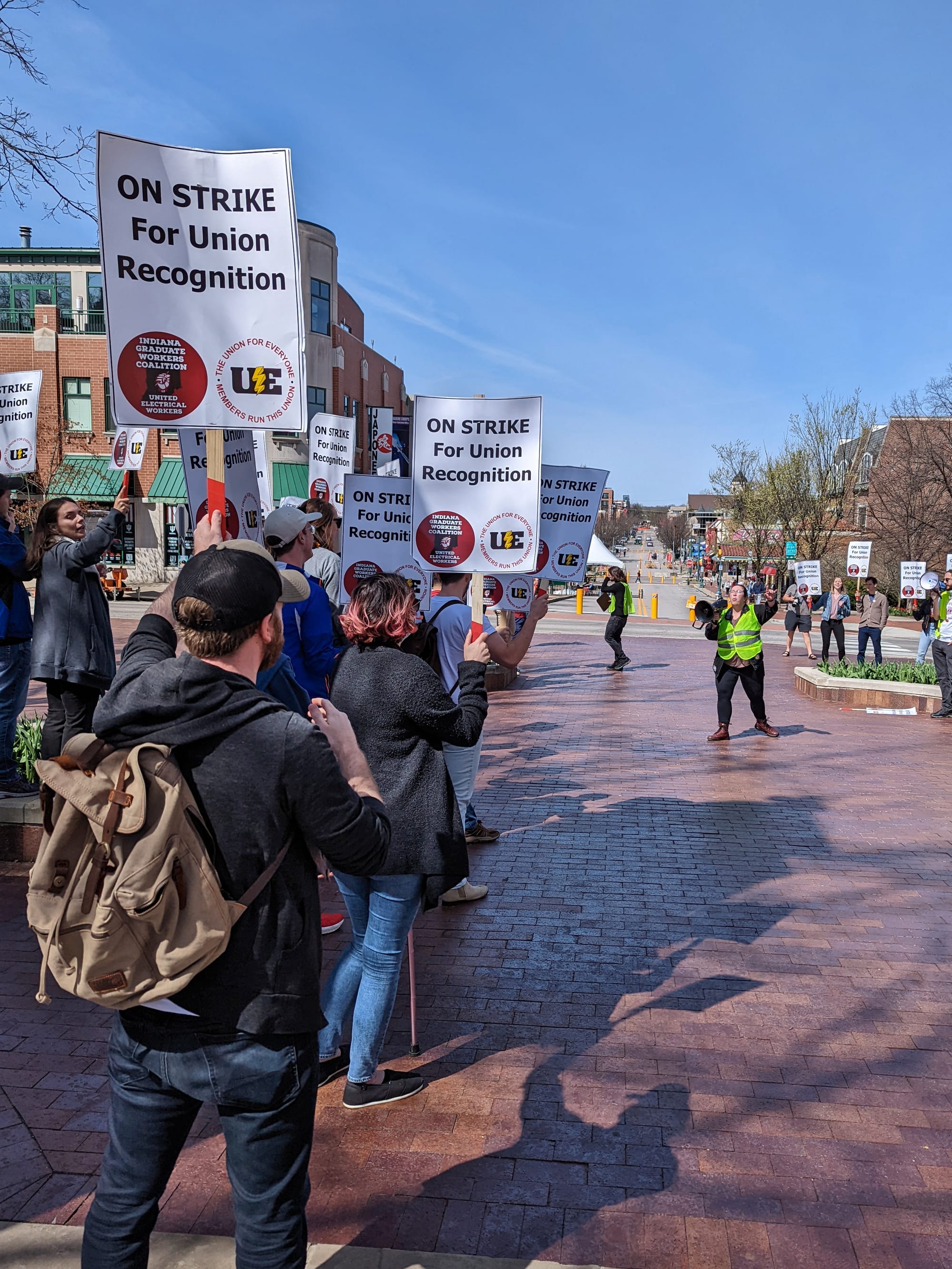 long shot down street with people on the left of the frame holding signs. Long shot of group of people standing in front of university gateway holding signs Woman in yellow reflective vest standing in the middle of a crowd of people with signs. The signs say:"On strike for union recognition."