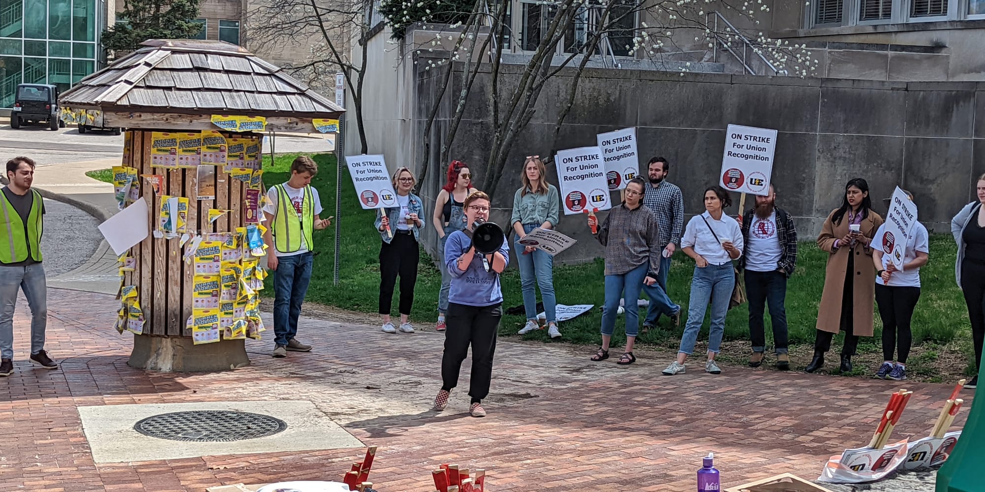 Woman speaking into a megaphone standing in the center of a group of people who are holding signs. The signs say "On strike for union recognition."