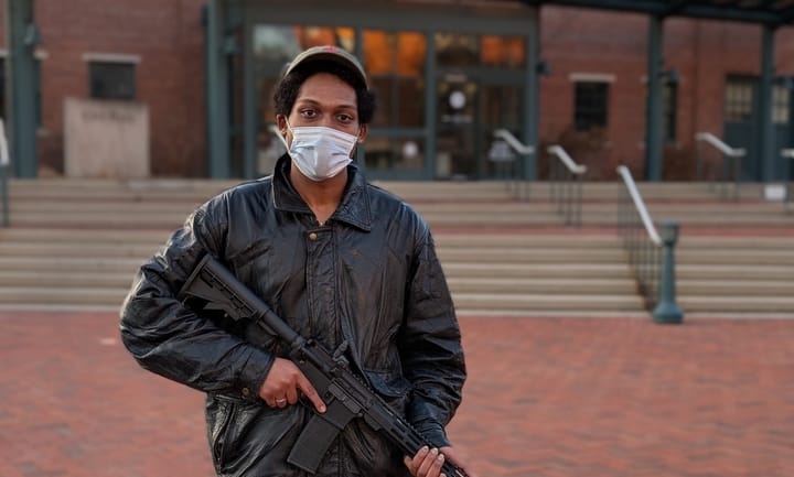 A person holds a military style rifle in front of a government building. The plaza is made of brick.