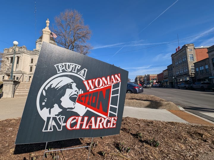 A sign placed in the flower bed says, "Put a woman in charge." A woman has a megaphone with the word "ACTION" written on it.